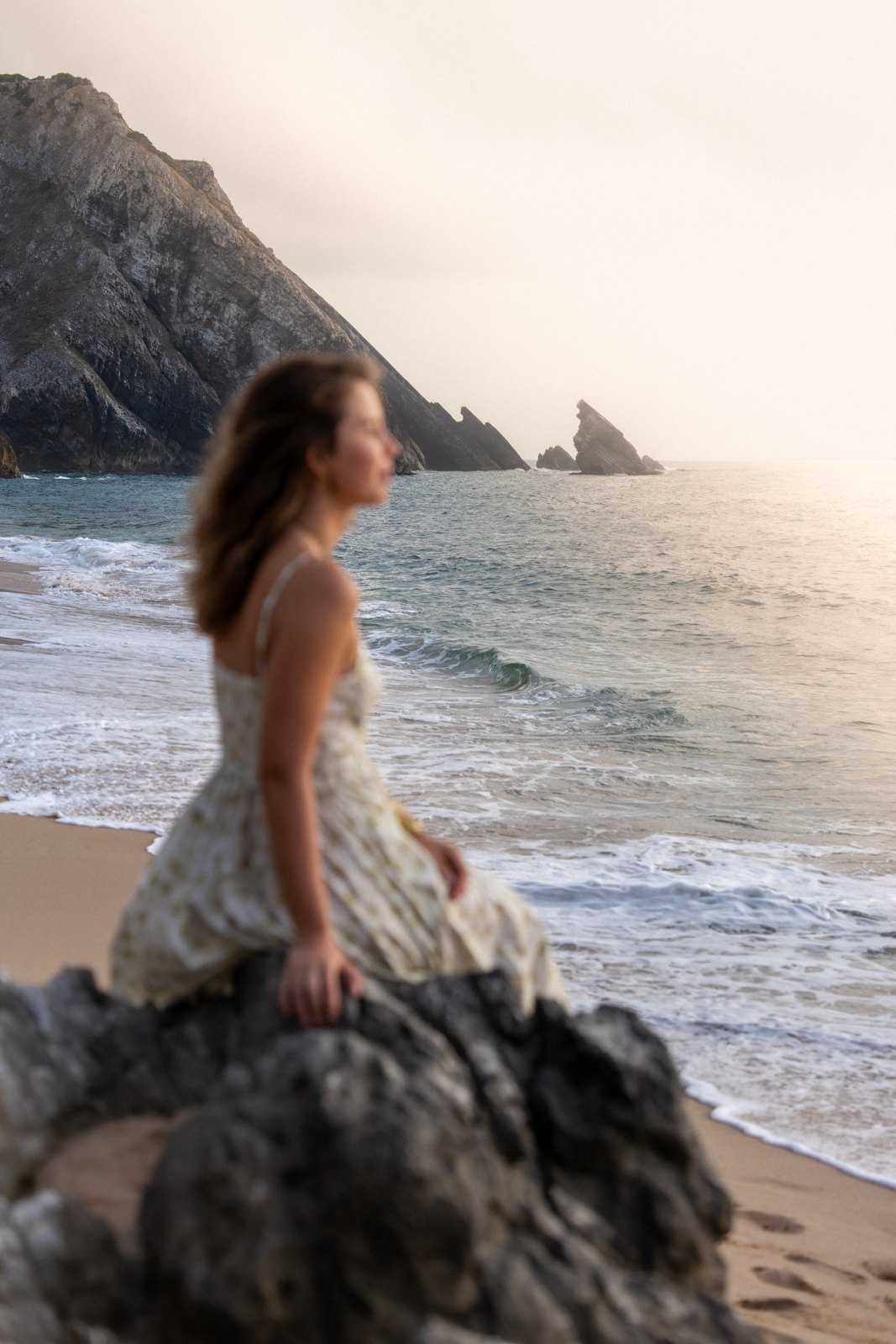 Woman in a light summer dress sitting on the rocks at Praia da Adraga, Sintra, at golden hour