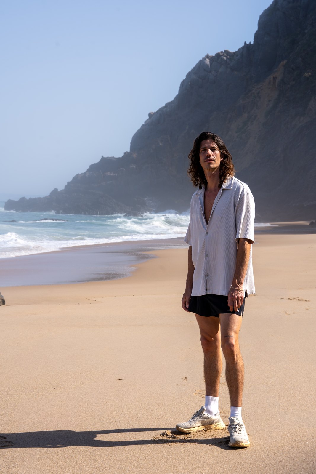 Man in an open shirt standing on a beach below dark cliffs