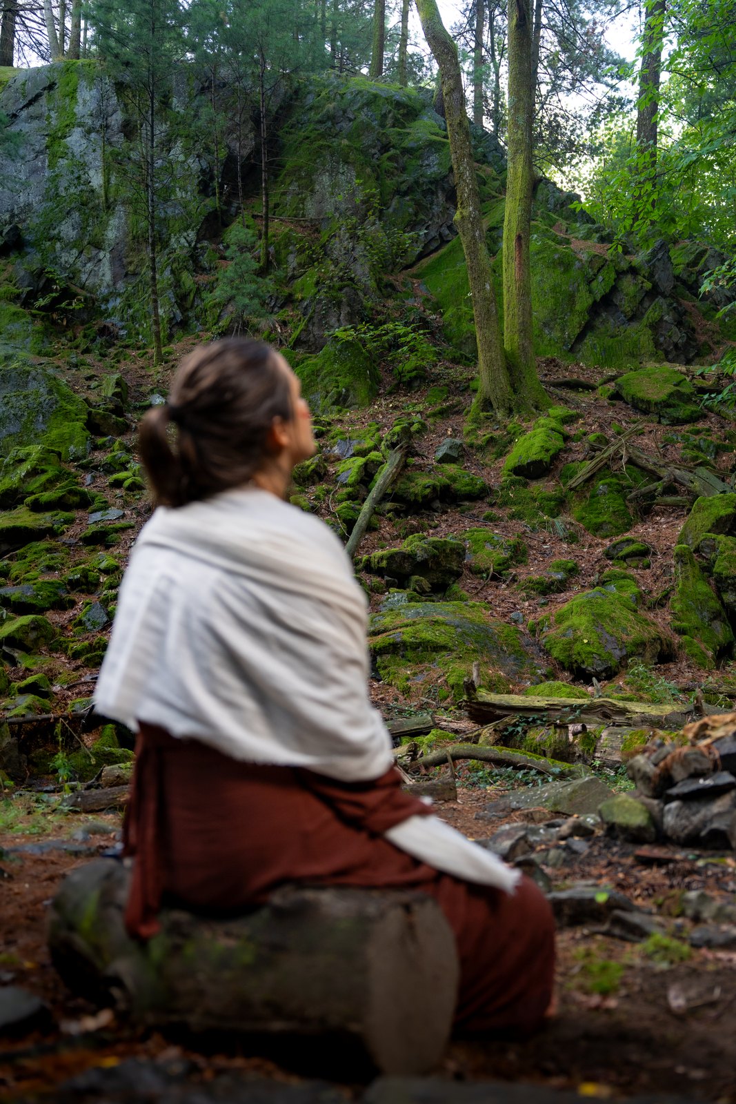 Woman in a white shawl sitting on mossy rocks in a forest