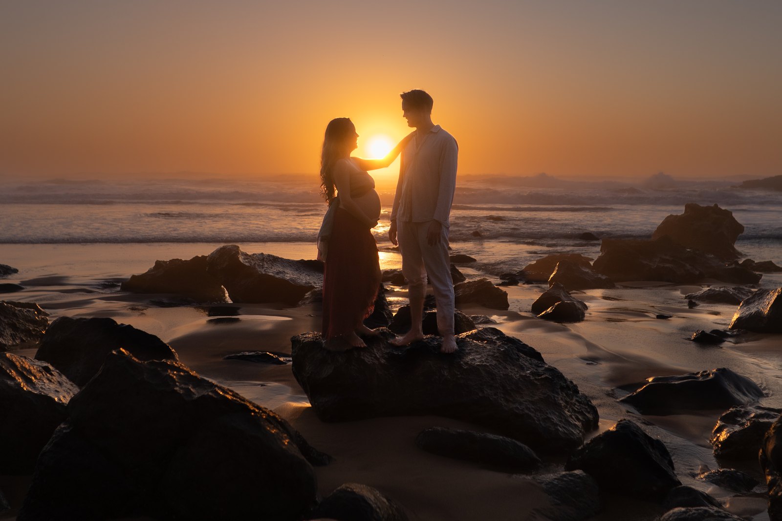 Pregnant couple standing on rocks by the sea at sunset