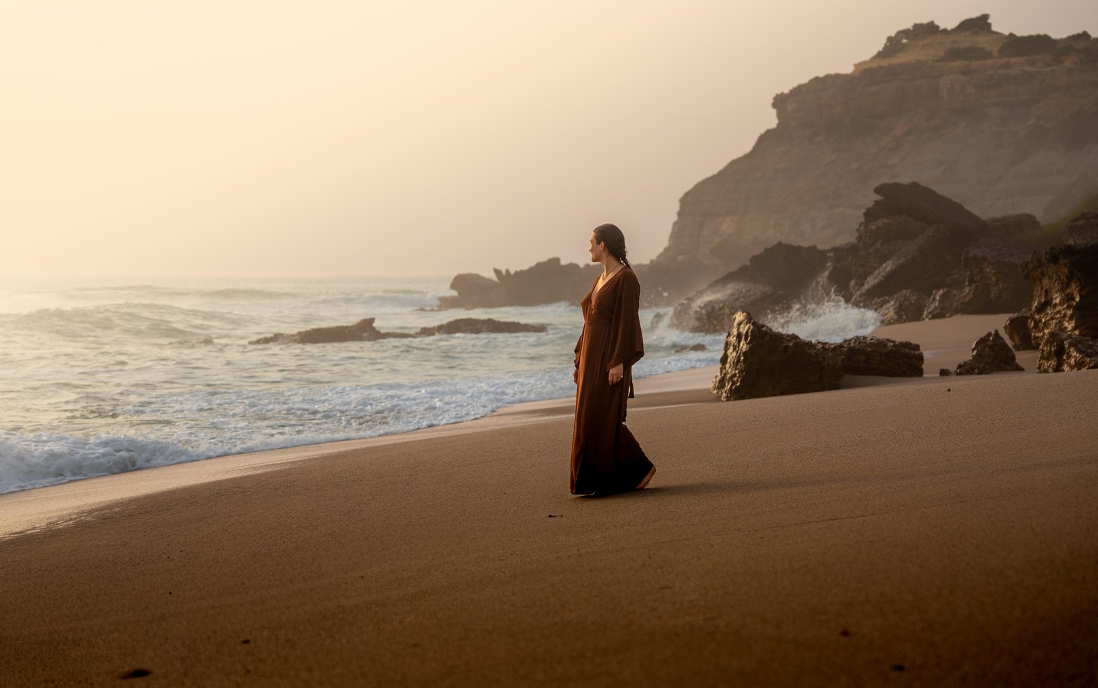 Woman in a long dress walking on a beach below cliffs