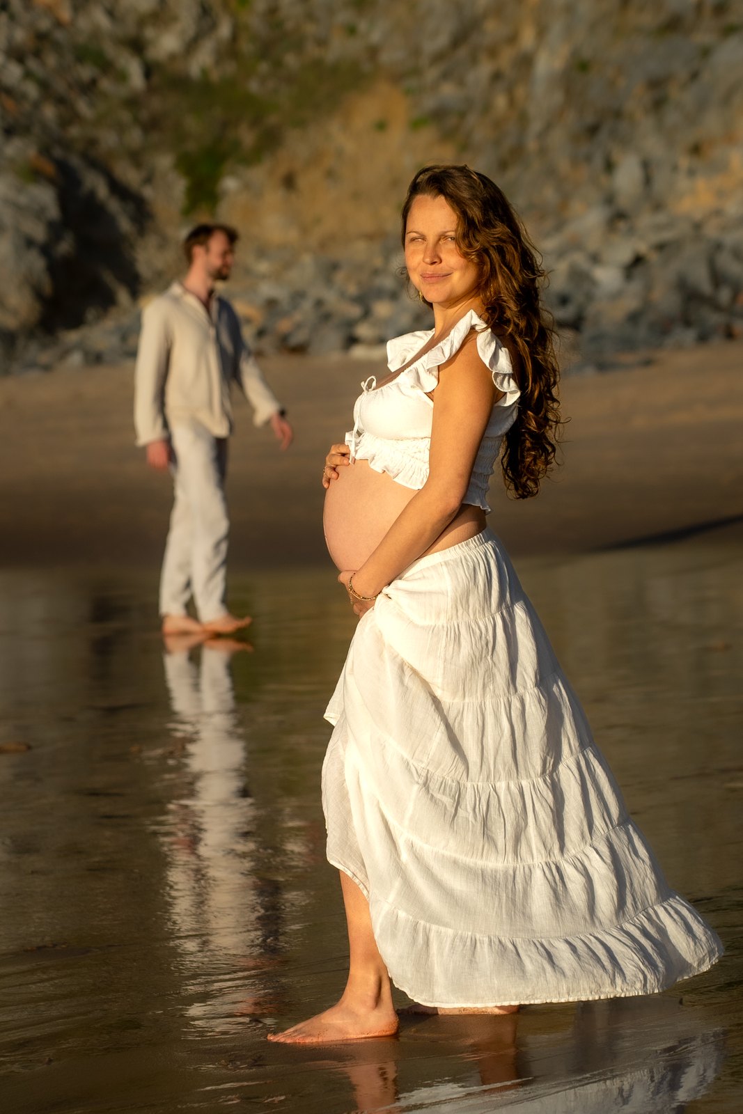 Pregnant woman smiling on the beach, her partner behind her